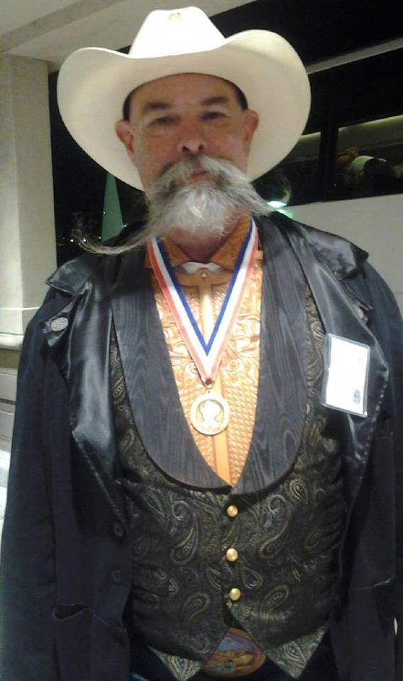 Wc wearing a white cowboy hat, a carved leather collar and bib under a black paisley vest, and black jacket with the Al Stohlman Award for Achievement in Leathercraft hung from a red white and blue ribbon around his neck
